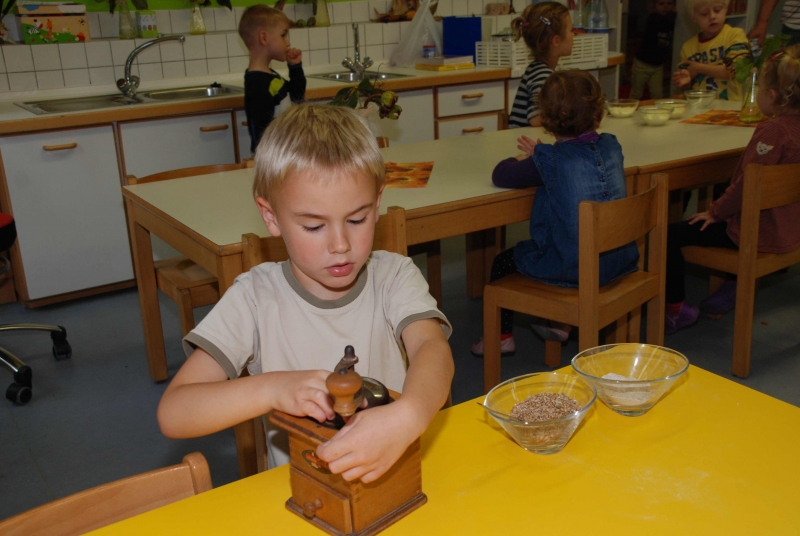 Engagiertes Arbeiten in der Küche. Im Vordergrund ein Junge, der mit einer Holzmühle Getreide mahlt.