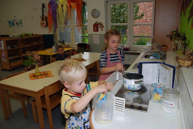 Zwei Kinder Kochen Pudding in der Küche. Am Tisch warten zwei weitere Kinder auf den Pudding.